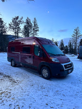A maroon Winnebago Solis RV rental is parked in a quiet, snowy winter forest under a crescent moon.