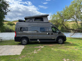Gray Winnebago Solis RV rental parked by a lake. This camper van with a pop-top roof is on grass.