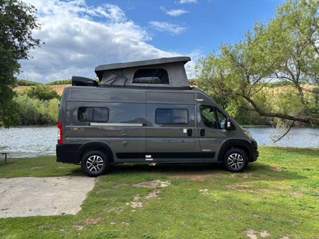 Gray Winnebago Solis RV rental parked by a lake. This camper van with a pop-top roof is on grass.