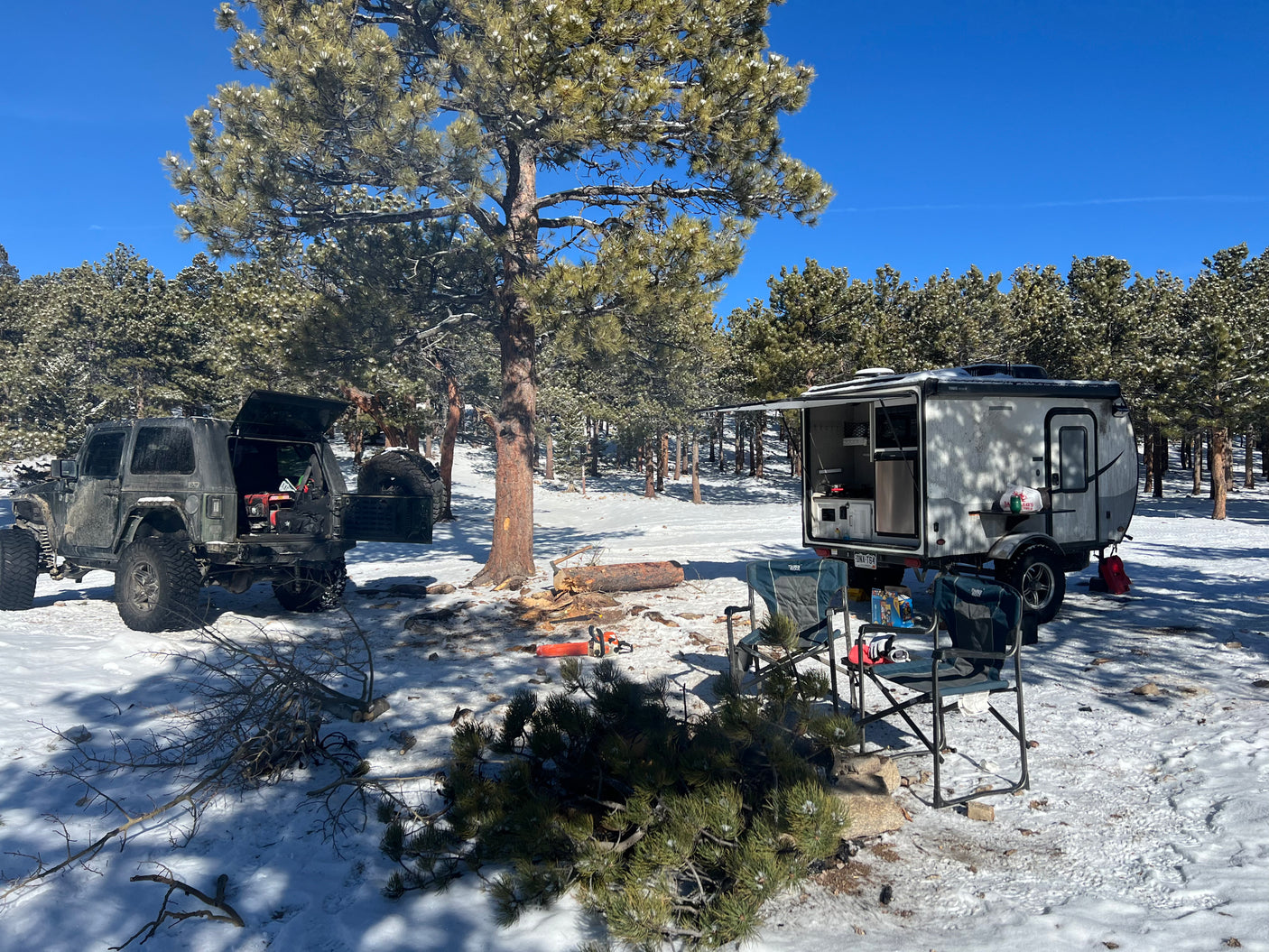 Winter camping scene with a rugged Jeep & RV rental. Preparing firewood near the small camper on snow-covered ground.