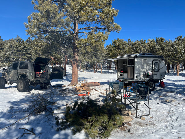 Winter camping scene with a rugged Jeep & RV rental. Preparing firewood near the small camper on snow-covered ground.