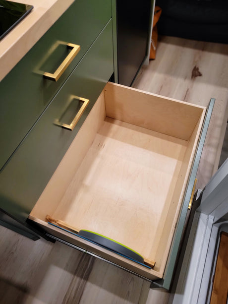 Inside an RV rental: a clean, open kitchen drawer made of light wood, part of stylish dark green cabinets with gold handles.
