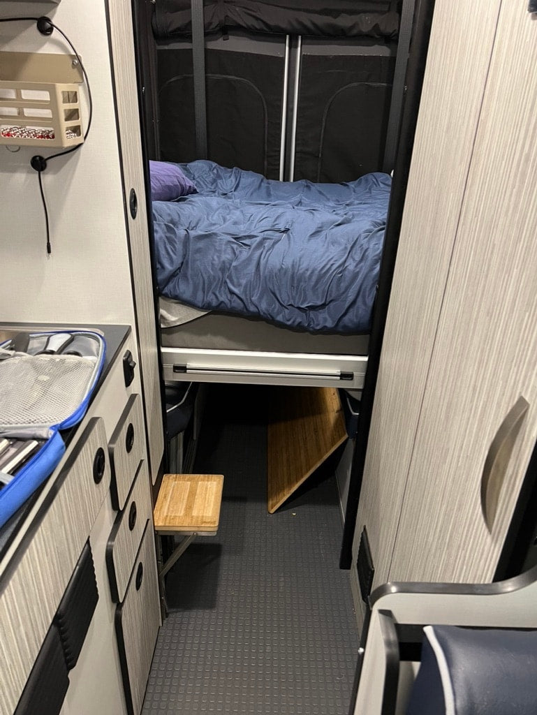  and a bike rack.|Open rear doors of an RV rental show a spacious interior with an elevated bed & storage for adventures.|Cozy RV rental bedroom interior with a comfortable bed