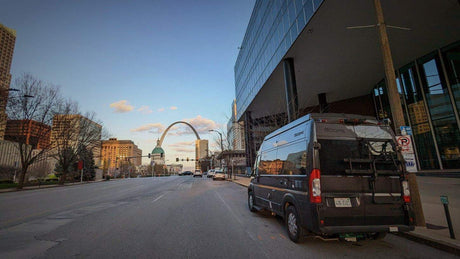 Black camper van RV rental parked on a city street with St. Louis's iconic Gateway Arch in the distance.