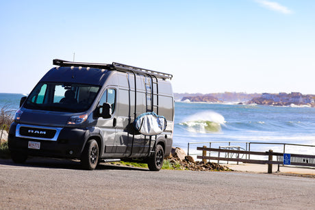 A dark gray camper van with a roof rack and surf bag overlooks crashing ocean waves. Ideal rv rental for beach adventures!