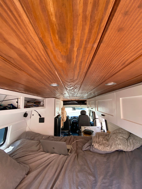 RV rental interior: warm wooden walls, white cabinets, cozy bed with laptop, and a compact kitchen.