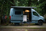 A sleek camper van RV rental with bikes ready for adventure in a tranquil forest campsite, side door open, illuminated inside.
