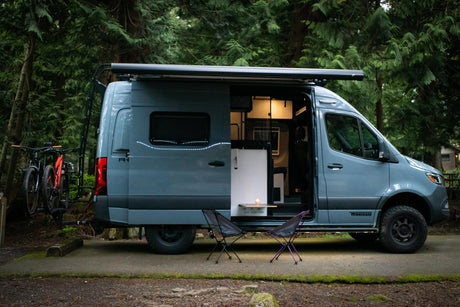 A sleek camper van RV rental with bikes ready for adventure in a tranquil forest campsite, side door open, illuminated inside.
