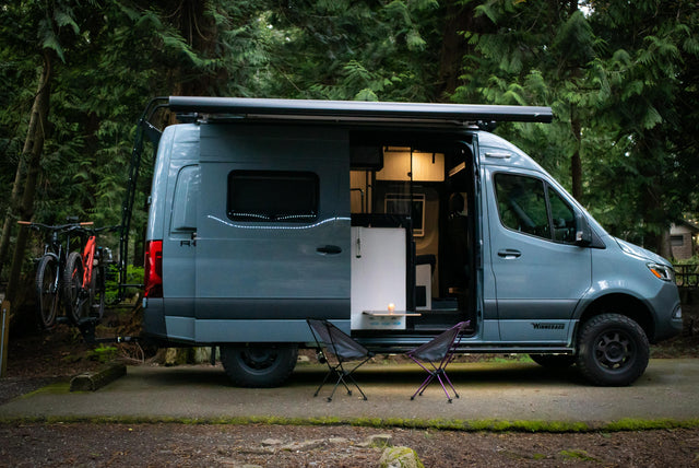 A sleek camper van RV rental with bikes ready for adventure in a tranquil forest campsite, side door open, illuminated inside.