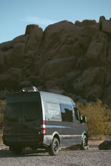 This 'The Tripsitter' Sprinter van, an RV rental, is parked on gravel near large desert rocks on a sunny day.