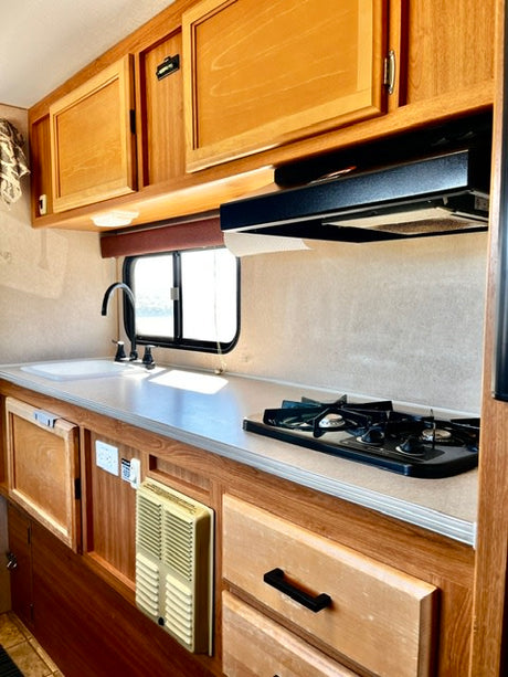 This rv rental kitchen has light wood cabinets, a sink, two-burner stovetop, black range hood, and a wall heater under a window.