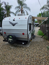 A retro-style travel trailer, a potential rv rental, is parked on gravel with its awning extended. Cloudy day.