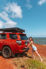 Red 4Runner SUV with an RV rental rooftop tent on a cliffside by the ocean. Woman smiles, ready for outdoor adventure.