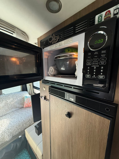 RV rental interior: A compact kitchen area featuring an open microwave with food, above a fridge, beside a cozy bed.