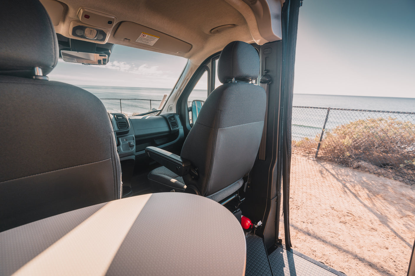 Inside an rv rental van, showing front seats, dashboard, & a coastal view with ocean, sand, & a fence. Fire extinguisher visible.