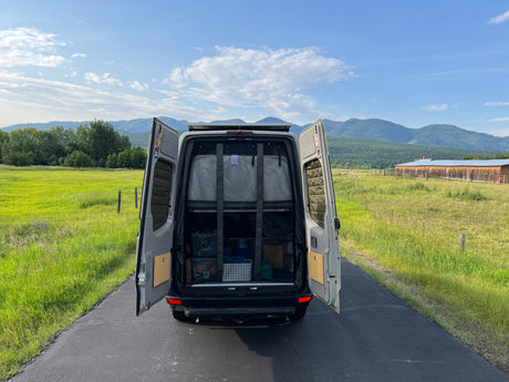 This rv rental van, with its rear doors open, showcases a bed and storage. Parked on a road in a scenic valley with green fields, mountains, and a barn.