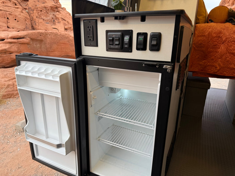 Inside an RV rental: open mini-fridge, control panel with switches/outlet, and red rock desert outside. — photo 18