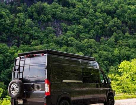 This sleek dark camper van, ideal for RV rental adventures, is parked amidst a vibrant green forest.