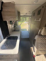 Tour this RV rental: compact kitchen with sink/stove, comfortable seating, and a scenic outdoor view through the open back.