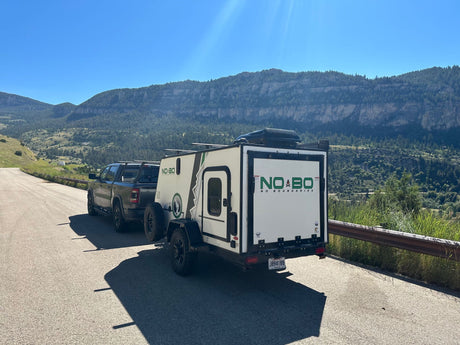 Truck tows a white camper trailer (RV rental) on a mountain road. Scenic outdoor adventure awaits under clear skies.