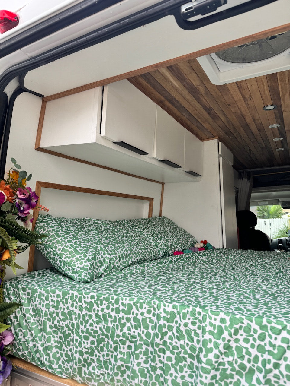 Cozy RV rental interior featuring a comfy bed with green sheets, white overhead cabinets, wood ceiling, and a headboard frame.