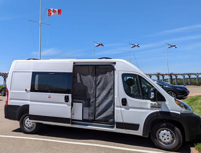 A white RV rental van with an open side door and screen is parked outdoors under Canadian and Nova Scotia flags.