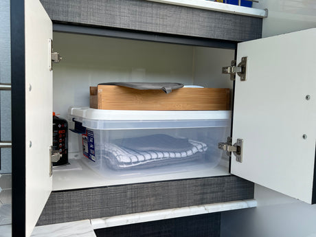 Practical RV rental storage: an open cabinet reveals neatly stacked clear containers with towels, a wooden tray, and a gas cylinder.