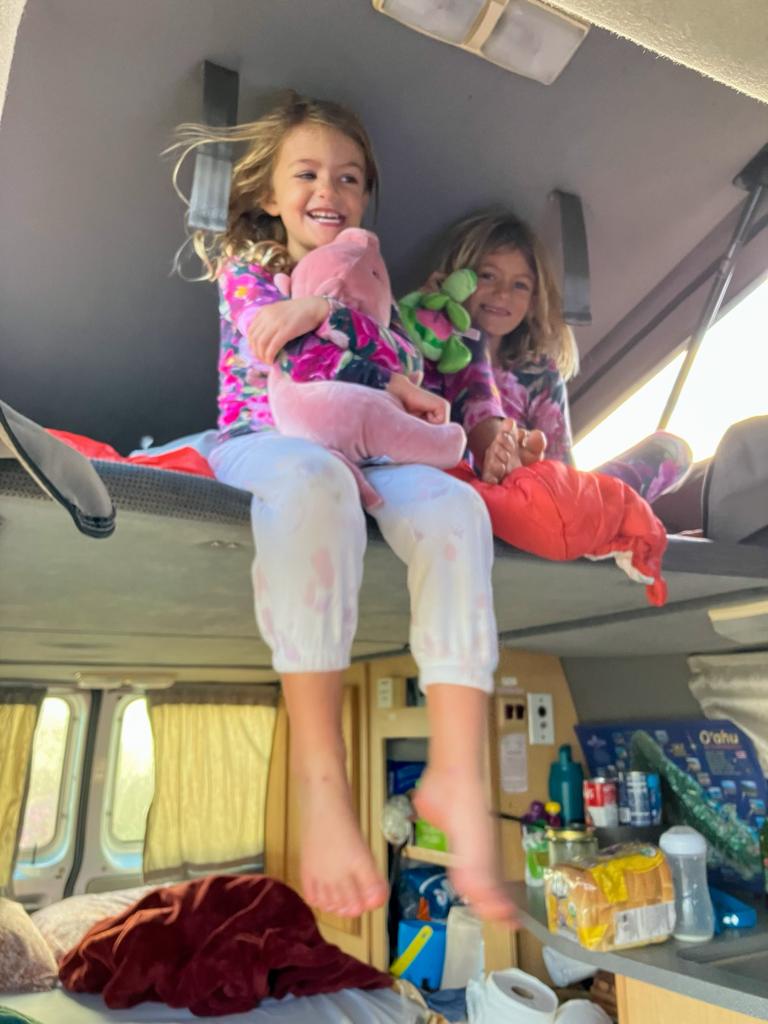 Two smiling girls in pajamas on an RV rental bunk bed, holding stuffed animals and enjoying the cozy van interior. — photo 3