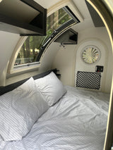 Cozy RV rental interior: a bed with striped white bedding, overhead storage, a large window revealing trees, and a compact design.