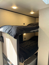 RV rental interior: two bunk beds with black comforters, warm overhead lighting, and light wood-paneled walls.