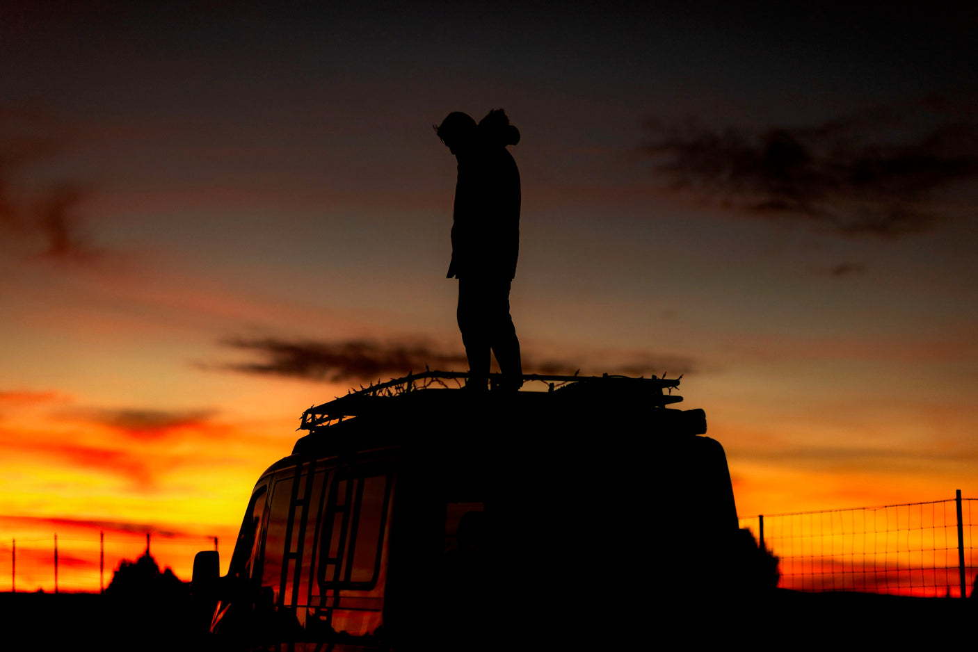 A person stands silhouetted on an RV rooftop against a vibrant orange and red sunset. Perfect for your next rv rental adventure.