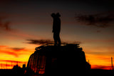 A person stands silhouetted on an RV rooftop against a vibrant orange and red sunset. Perfect for your next rv rental adventure.