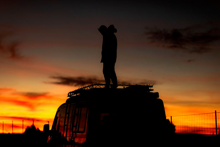 A person stands silhouetted on an RV rooftop against a vibrant orange and red sunset. Perfect for your next rv rental adventure.