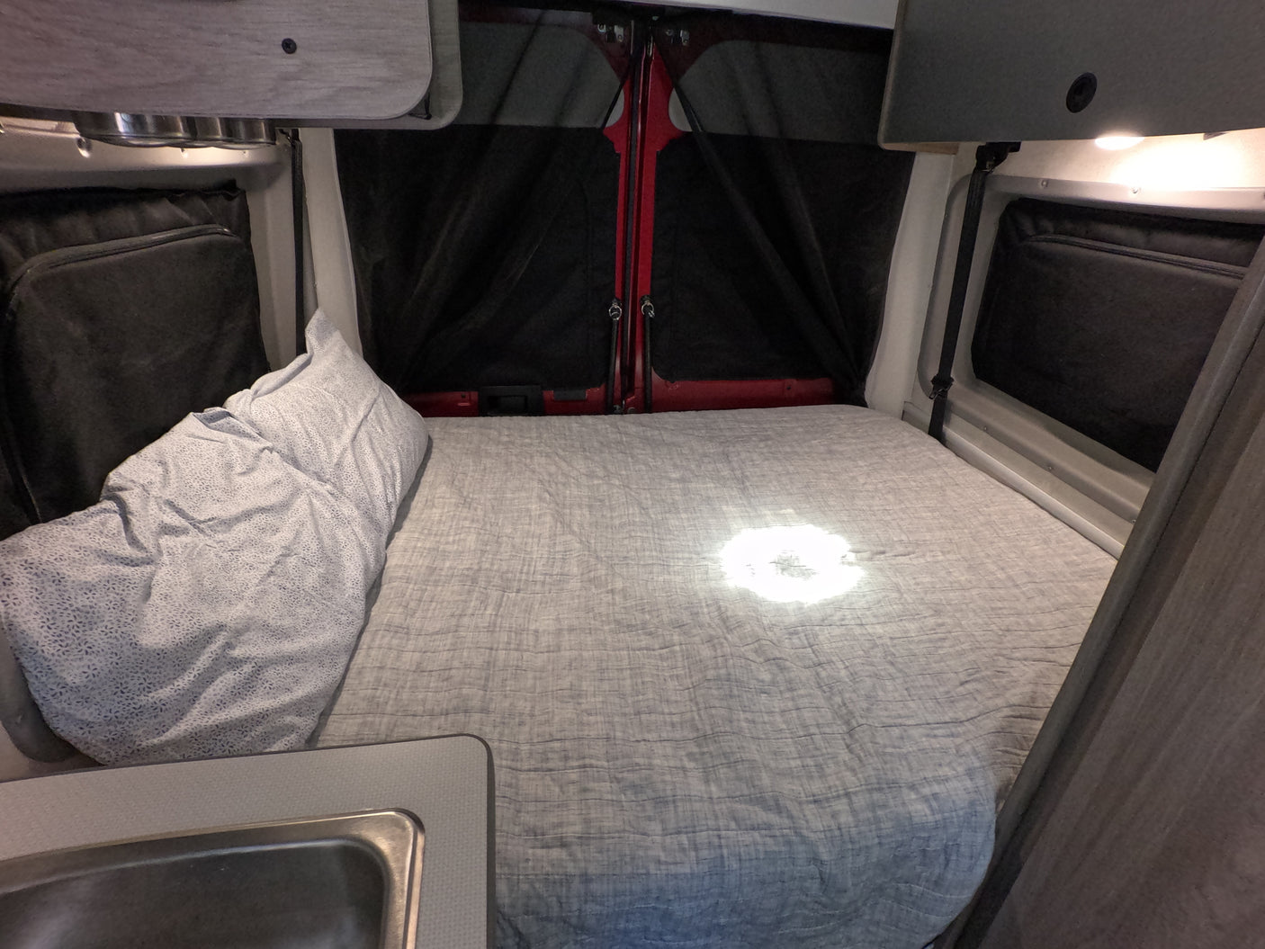 Inside an RV rental: a compact sleeping area with a comfortable bed, sink, and rear door screens for ventilation.