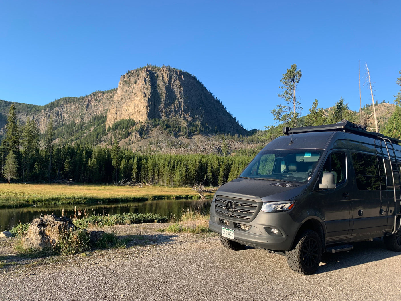A sleek Mercedes Sprinter RV rental is parked by a serene river, backed by majestic mountains and a lush forest.
