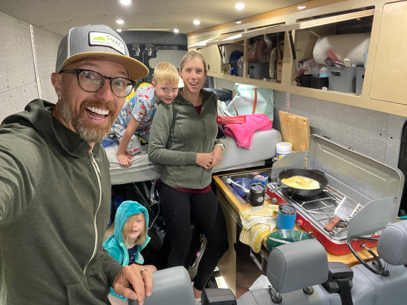 Family inside their RV rental: Dad's selfie, mom, and kids near bunk and stove cooking eggs for breakfast.