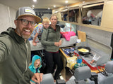 Family inside their RV rental: Dad's selfie, mom, and kids near bunk and stove cooking eggs for breakfast.
