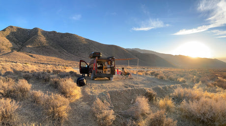Enjoying a desert sunset in an RV rental. A person relaxes outside their camper van with an awning, surrounded by mountains.
