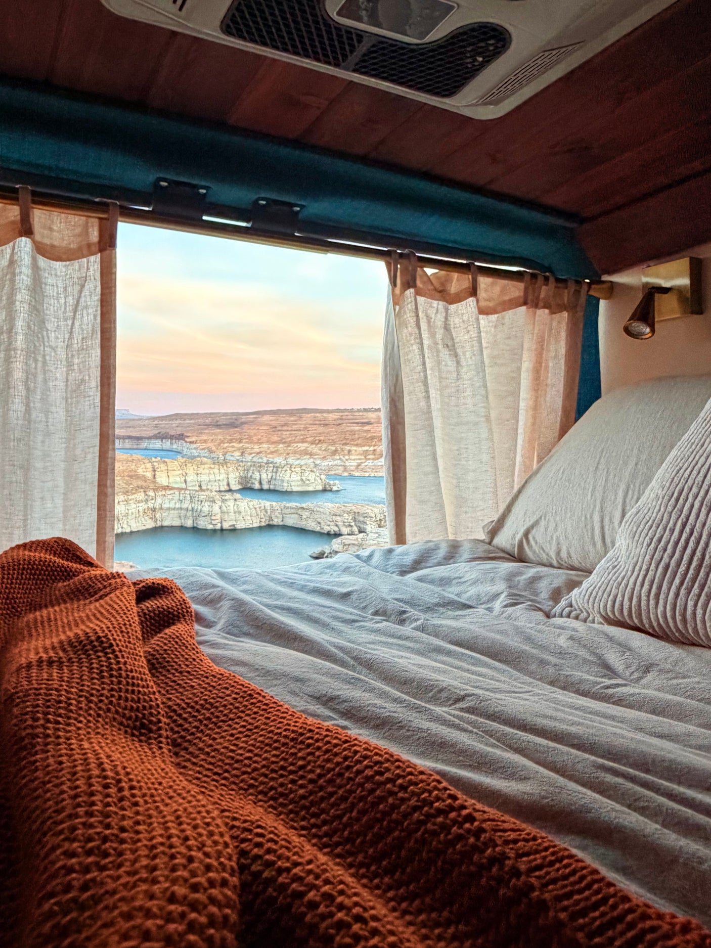 Relax in this cozy RV rental bed, offering expansive views of a lake nestled within rocky canyons at sunrise/sunset.