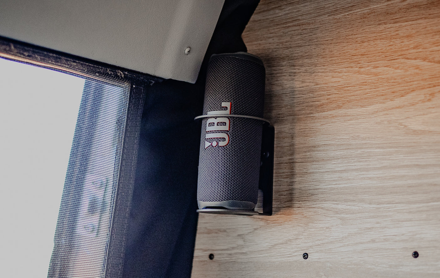 JBL portable speaker in a custom mount on wood panel, next to a window screen in an RV rental for easy tunes.