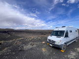 A white RV rental is parked on dark, rocky terrain, overlooking a massive volcanic crater with distant mountains under a cloudy sky.