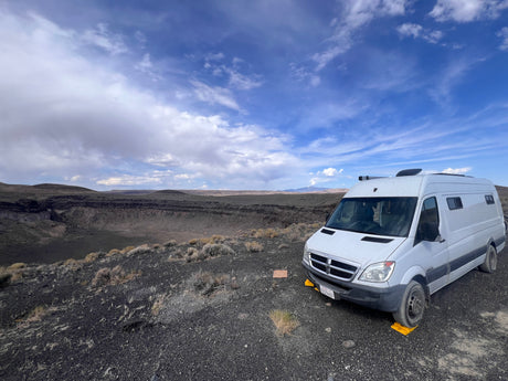 A white RV rental is parked on dark, rocky terrain, overlooking a massive volcanic crater with distant mountains under a cloudy sky.
