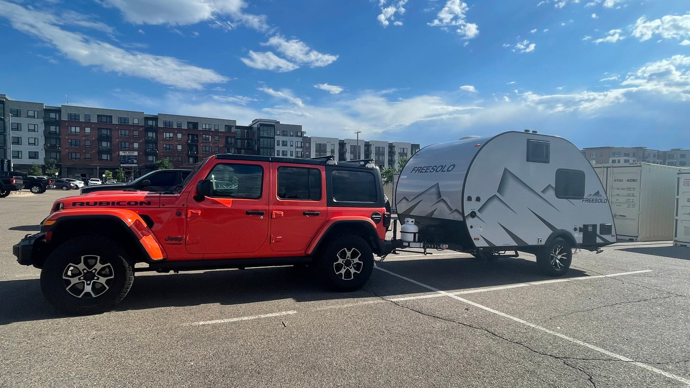 An orange Jeep Rubicon tows a Freesolo teardrop RV rental in a sunny parking lot, ready for travel.