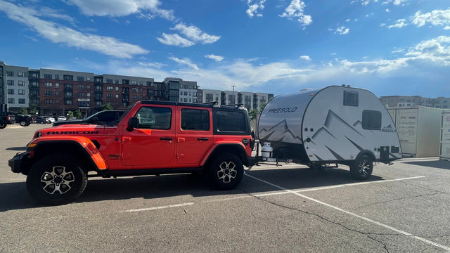 An orange Jeep Rubicon tows a Freesolo teardrop RV rental in a sunny parking lot, ready for travel.