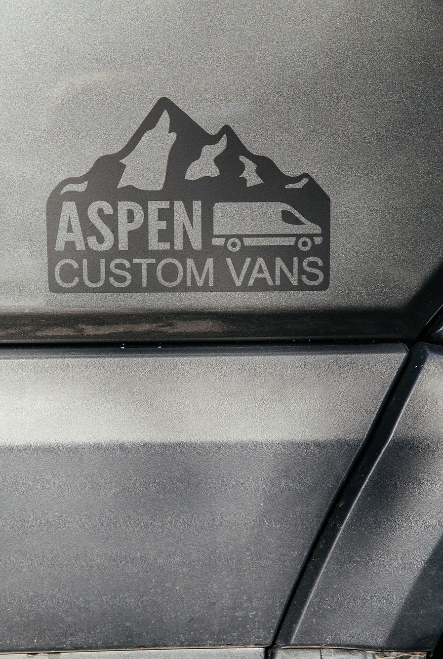 Aspen Custom Vans logo on a dark gray textured vehicle surface, featuring mountains & a camper van; great for rv rental.