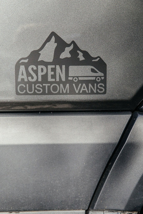 Aspen Custom Vans logo on a dark gray textured vehicle surface, featuring mountains & a camper van; great for rv rental.