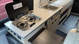 This RV rental interior shows a compact, light-toned kitchen with a two-burner gas stove and sink, ideal for camping adventures.
