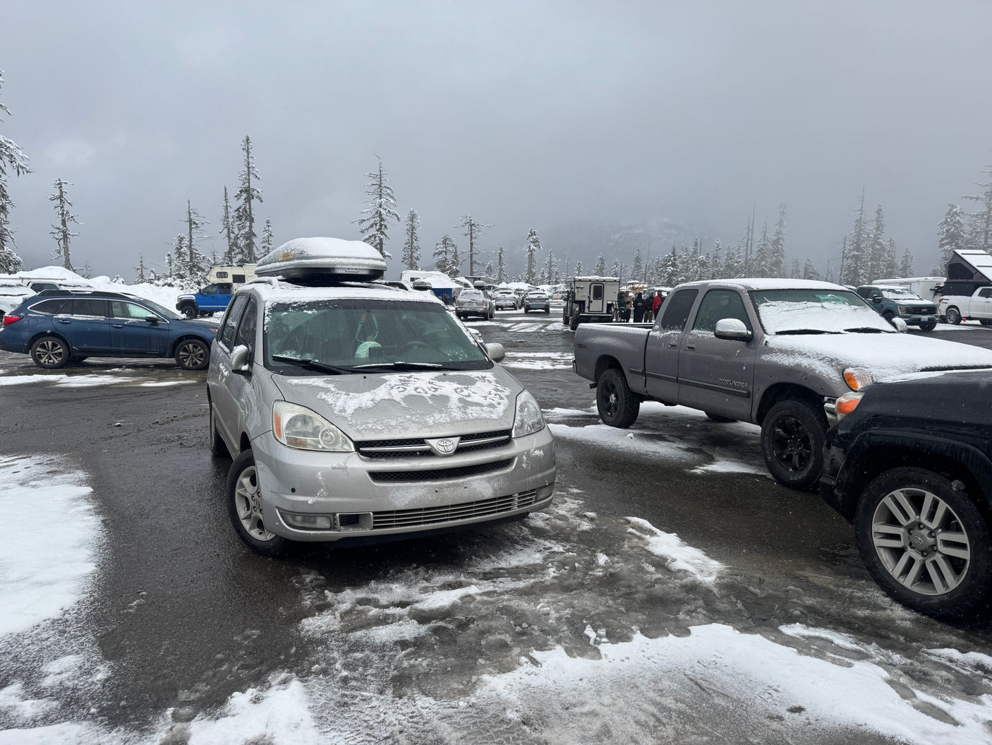 Snowy winter parking lot with cars, trucks, and an RV rental. Snow-covered trees under an overcast sky.