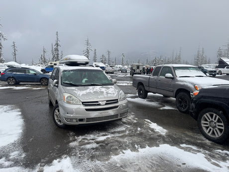 Snowy winter parking lot with cars, trucks, and an RV rental. Snow-covered trees under an overcast sky.