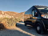 Black camper van RV rental parked on a desert road. Rocky mountains and clear sky reflected
